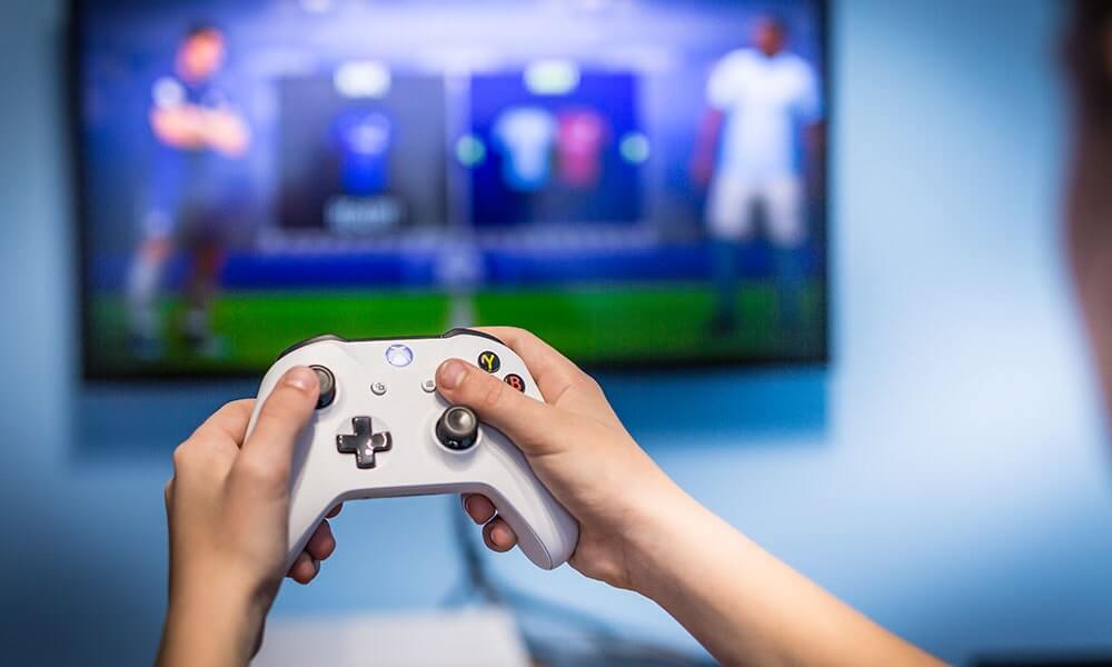 Hands holding a white Xbox One controller with a blurred monitor and game in the background